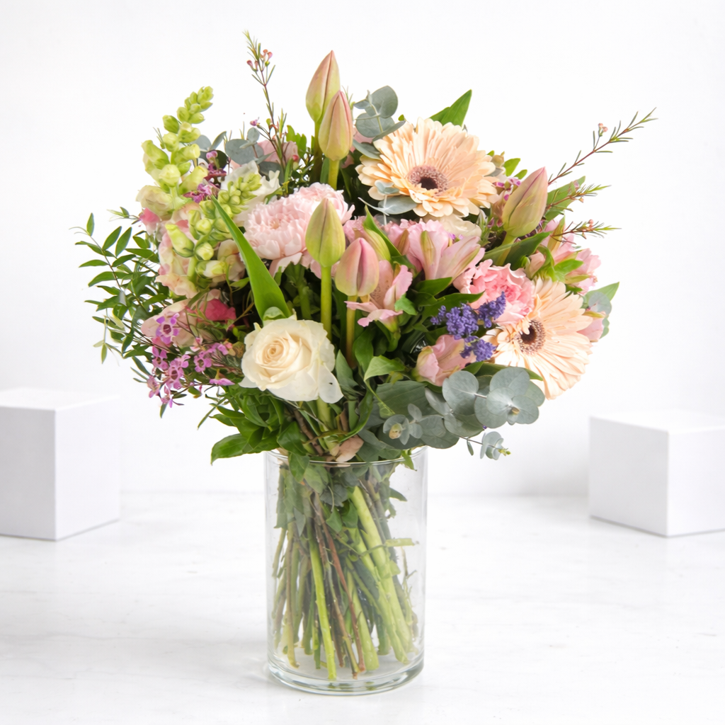 Soft pastel bouquet with peach gerberas, pink tulips, white roses, lavender, and eucalyptus in a clear glass vase on a white surface.