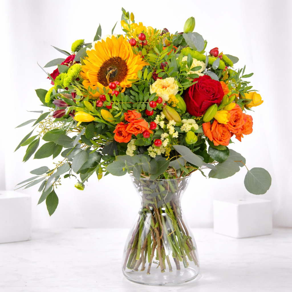 Vibrant bouquet with sunflower, red and orange roses, yellow tulips, red berries, and eucalyptus in a glass vase on a white surface.