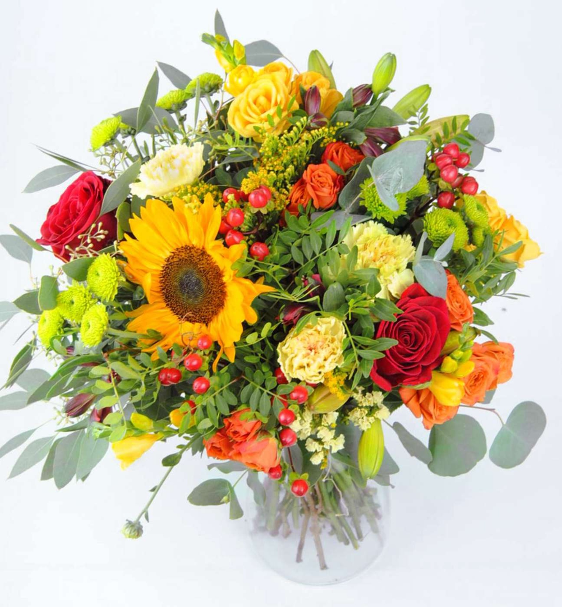 Top-down view of a vibrant bouquet with sunflower, red and orange roses, yellow carnations, red berries, and eucalyptus in a glass vase.