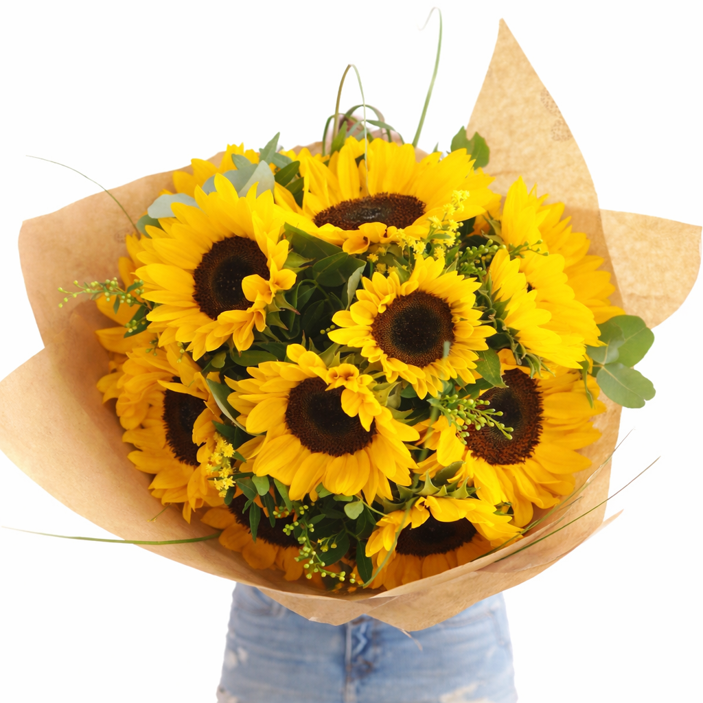 Person holding a large sunflower bouquet wrapped in kraft paper with green foliage accents.