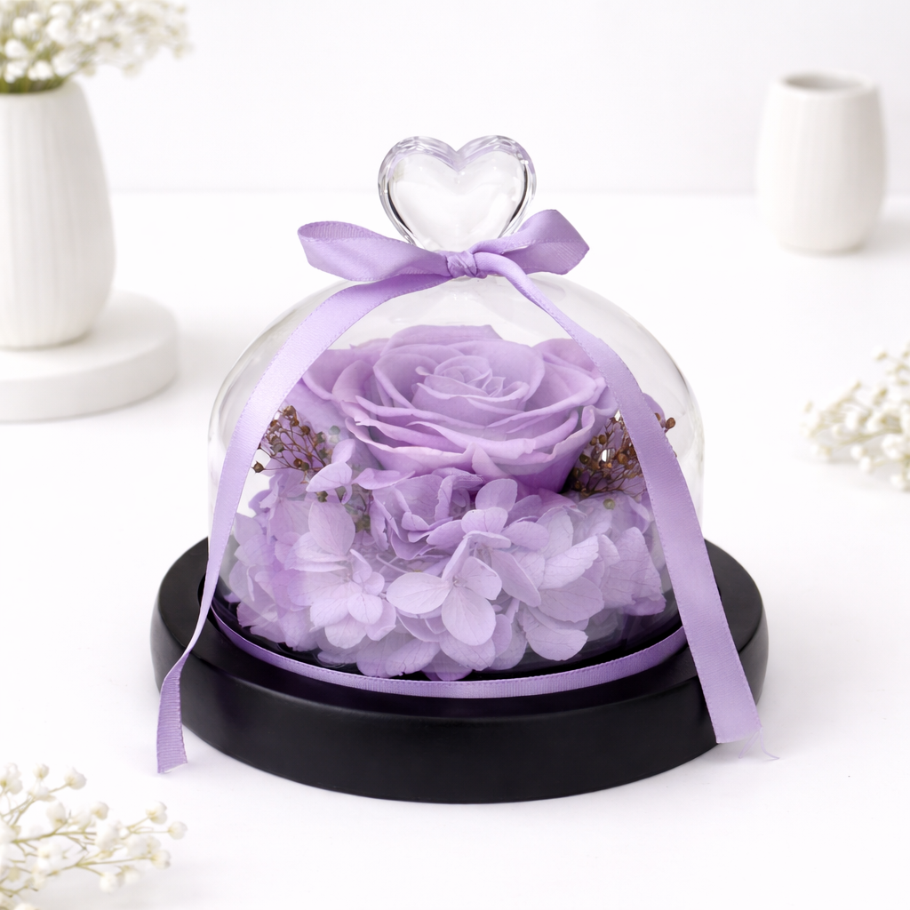 Purple preserved rose and hydrangeas enclosed in a glass dome with a crystal heart handle and purple ribbon bow on a black base.