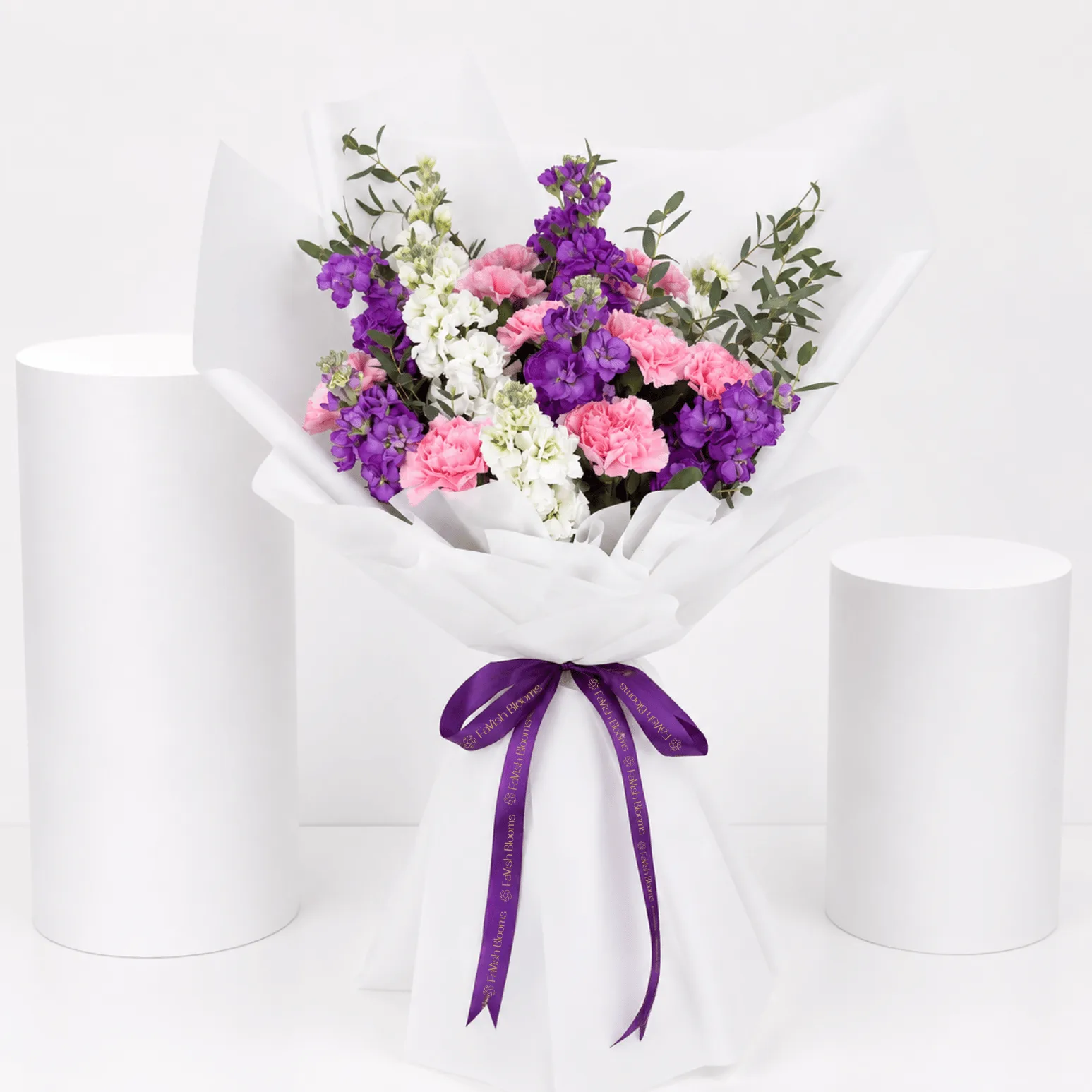Pastel bouquet with pink carnations, purple stock flowers, and white blooms wrapped in elegant white paper.