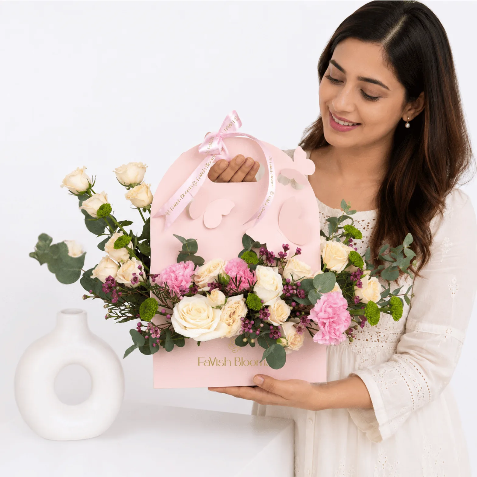 Smiling woman holding a pastel floral arrangement in a pink Favish Blooms gift bag featuring cream roses, pink carnations, eucalyptus, and seasonal blooms.